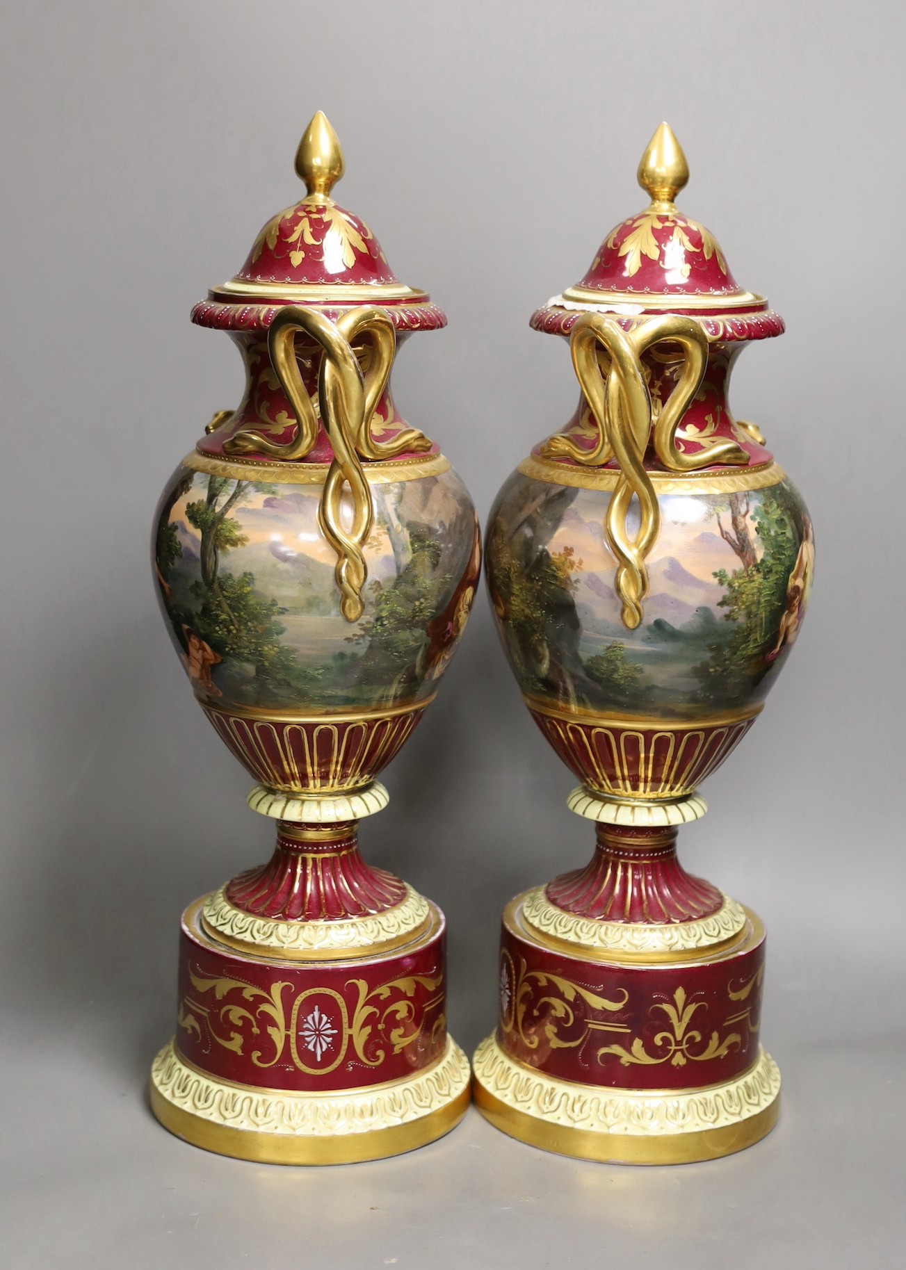 A pair of ornate Vienna style two handled urns bearing covers, with painted love scene and red ground and gilt decoration, 46.5cm high